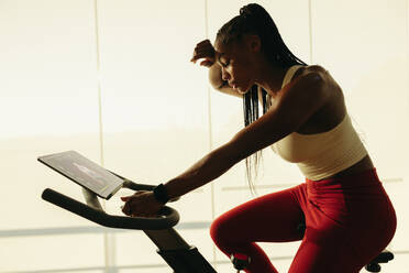 Young black woman sweating it out while working out with a cycling app. With her tablet nearby, she follows the online workout routine, pushing herself to achieve her fitness goals. - JLPSF30522