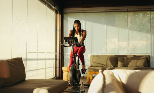 Woman doing a cardio workout on a stationary bicycle in her home, as part of her routine for a healthy lifestyle. She pedals with determination, striving to achieve her fitness goals and improve her health. - JLPSF30523