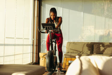 Black woman achieves her fitness goals with the help of an online cycling class. With her tablet in front of her, she follows along with the virtual workout routine with focus and determination. - JLPSF30524