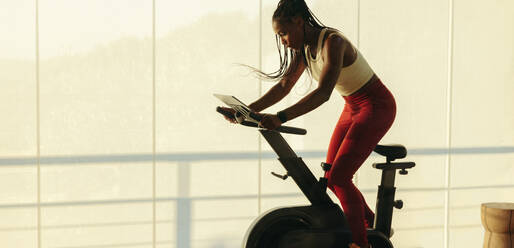 Young woman getting into her home workout routine, using a smart exercise bike and a fitness app for indoor cycling. Black female using high-tech fitness equipment to stay fit and healthy. - JLPSF30528