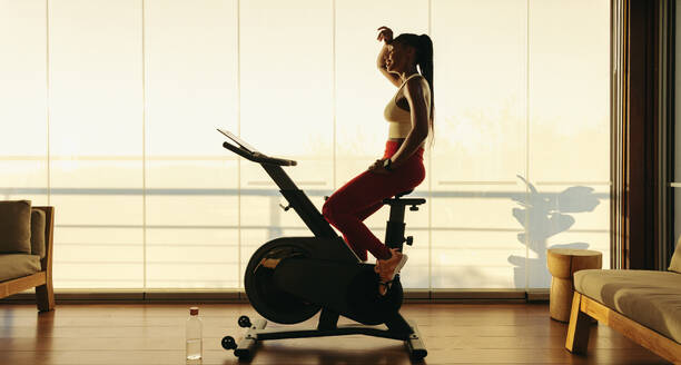 Woman works out with a smart stationary bike at home, she takes a break from exercising and rests, wiping off her sweat. She is determined to continue her fitness training and maintain a healthy lifestyle. - JLPSF30533