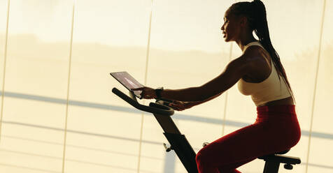 Young African woman exercising on a smart exercise bike with a touchscreen tablet. She's participating in a digital fitness class at home, working towards her fitness goals through virtual training. - JLPSF30535