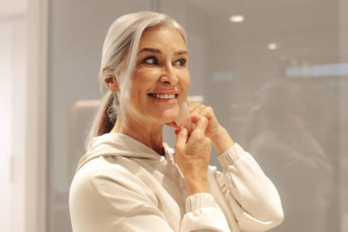 Senior woman massages her face with a gua sha stone as part of her skincare routine. With a serene and focused expression on her face, she enjoys the self-care ritual and the benefits it provides for her skin. - JLPSF30545