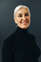 Portrait of a happy senior woman with silver hair smiles at the camera as she stands in a studio, expressing confidence in herself as a grey-haired female professional. - JLPSF30555