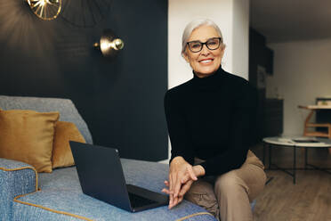 Senior female freelancer balances work and life as she sits comfortably on a couch with a laptop, looking at the camera with a smile. Happy mature woman working from home. - JLPSF30561