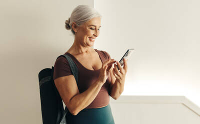 Senior woman holding a smartphone with a smile on her face, using an online fitness app to guide her yoga routine at home. Mature woman embracing a healthy lifestyle as she ages. - JLPSF30565