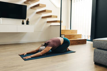 Mature woman practicing balasana, the child’s pose in yoga, in the comfort of her home. Senior woman embracing a healthy lifestyle in her old age, grounding herself and finding inner calm. - JLPSF30570