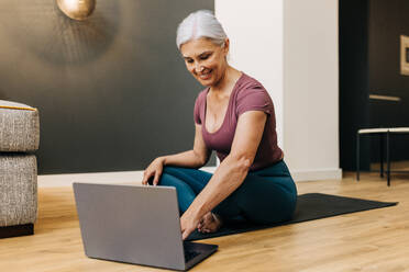 Senior woman uses her laptop to discover and explore new yoga classes, exploring new routines of wellness and self-care. Caucasian woman seeking to deepen her fitness practice and improve her health. - JLPSF30578