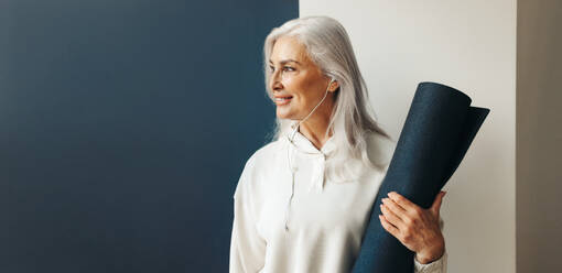 Senior woman stands holding a yoga mat, with a peaceful expression on her face. Mature woman preparing to begin her yoga practice, part of her fitness and self-care journey as an aging woman. - JLPSF30581