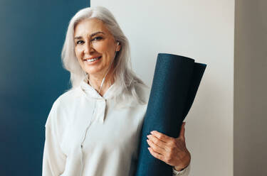 Mature woman stands holding a rolled-up yoga mat, preparing to start her yoga practice for the day. Senior woman embracing a healthy lifestyle with the addition of yoga to her fitness routine. - JLPSF30582