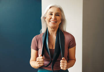 Happy senior woman stands holding resistance bands around her neck, with a joyful smile on her face. She is dressed in workout clothes and appears to be embracing the fitness lifestyle, ready to challenge herself and achieve her fitness goals. - JLPSF30583
