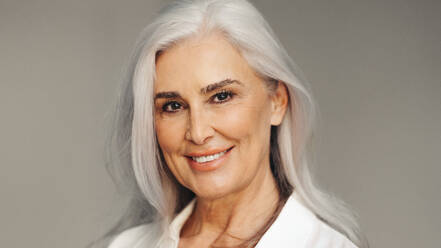 Professional business woman with silver hair stands confidently against a grey background, smiling warmly at the camera. She embodies a sense of confidence and competence, demonstrating her professionalism and experience. - JLPSF30588