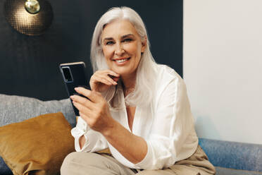 Happy senior woman sits on a cozy couch, using her smartphone to stay connected in her retirement. Woman using a smart mobile device to keep herself engaged in her leisure time. - JLPSF30590