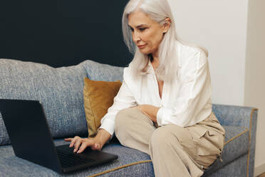 Senior woman working as a freelancer, using her laptop to work from the comfort of her home. Sitting on a couch, she balances work and relaxation as she communicates with clients, writes reports, and manages her projects. - JLPSF30591