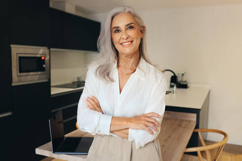 Mature real estate agent standing in a house, smiling at the camera. With her laptop on the table behind her, she is ready to sell a beautiful furnished home. - JLPSF30592