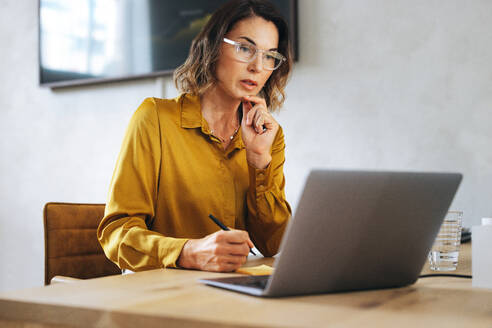 Professional business woman participates in an online meeting from her office. With her laptop and sticky notes, she engages with colleagues through video conferencing, discussing an important project. - JLPPF02051