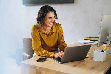 Happy and accomplished businesswoman sits in her office, typing on her laptop with a smile. Successful web designer utilizing her expertise and creativity to achieve success in her business ventures. - JLPPF02053