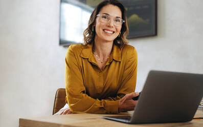 Portrait of a Caucasian businesswoman, wearing eyeglasses and sitting in her office. Professional woman smiling at the camera, representing the achievements and accomplishments of a dedicated professional. - JLPPF02064