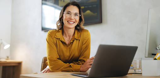 Successful businesswoman with eyeglasses looking at the camera with a warm and genuine smile. Woman sitting at her office desk portraying hard work, success and entrepreneurship. - JLPPF02065