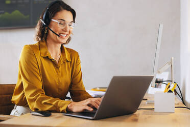 Happy call center agent using a laptop with a headset on. Woman assisting customers with technical support at her office desk, answering their calls and providing solutions through her microphone. - JLPPF02076