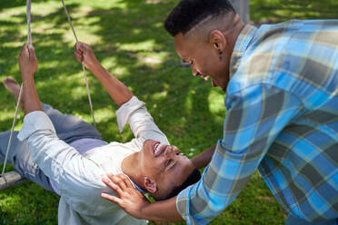 Happy young gay male couple on swing in summer park - CAIF33419