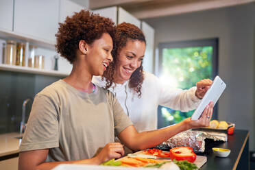 Happy lesbian couple with digital tablet cooking, looking at recipe - CAIF33465