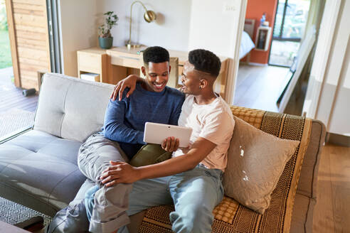 Happy young gay male couple hugging and using digital tablet on sofa - CAIF33467