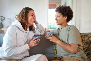 Happy lesbian couple drinking coffee on living room sofa at home - CAIF33473