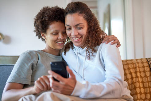 Happy lesbian couple hugging, using smart phone on sofa at home - CAIF33482