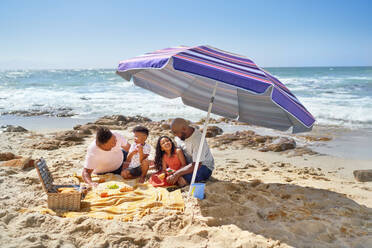 Gay male couple and kids eating lunch under umbrella on sunny beach - CAIF33523