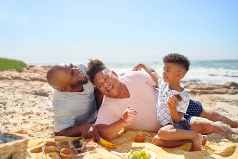 Happy gay male couple and son laughing and eating on sunny beach - CAIF33525