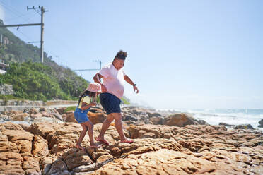Barefoot father and daughter walking on rocks on sunny beach - CAIF33529