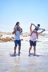 Gay male couple carrying kids on shoulders sunny ocean surf - CAIF33536