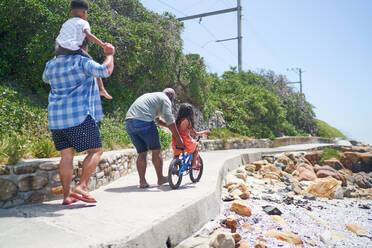 Gay male couple with kids riding bike on sunny beach boardwalk - CAIF33546