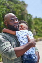 Affectionate father holding tired son on sunny footpath - CAIF33547