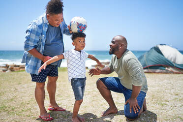 Gay male couple and son playing with soccer ball on sunny beach - CAIF33550