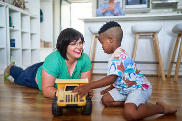 Happy mother and son playing with toy dump truck on floor at home - CAIF33578