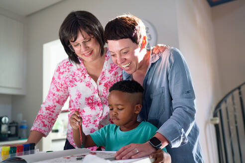 Happy lesbian couple watching son painting in kitchen at home - CAIF33593