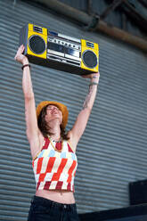 Carefree young woman holding boom box overhead in alley - CAIF33657