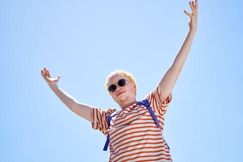 Portrait cool young man with arms raised below sunny blue sky - CAIF33728