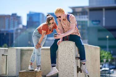 Young friends using smart phone at ledge in sunny city - CAIF33730