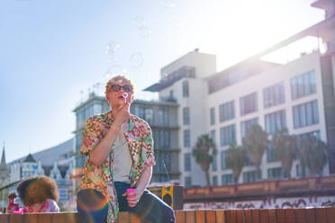 Young man blowing bubbles on sunny city urban balcony - CAIF33765