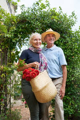 Happy senior couple with flowers walking under trellis in garden - CAIF33809