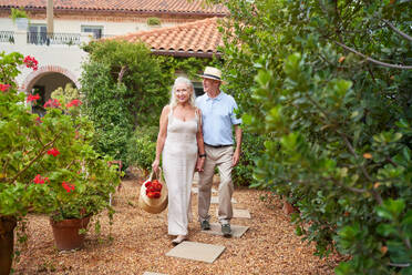 Happy senior couple with flowers walking in garden outside villa - CAIF33814