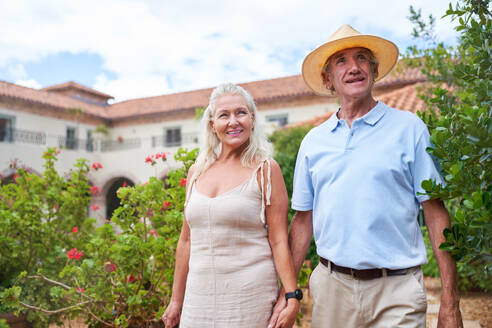 Portrait happy senior couple walking in garden outside villa - CAIF33815
