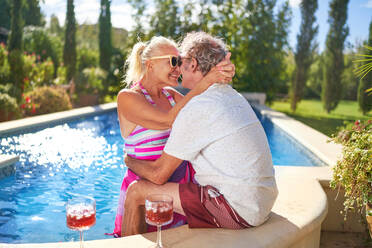 Happy senior couple hugging and kissing at sunny swimming pool - CAIF33866
