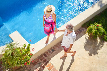 Senior couple holding hands, walking along summer swimming pool - CAIF33874
