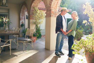 Happy senior couple holding hands on sunny villa patio - CAIF33881