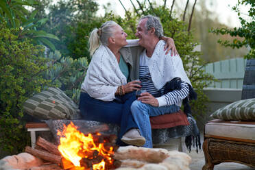 Happy senior couple drinking wine and hugging by fire pit on patio - CAIF33902