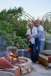 Happy senior couple hugging by fire pit on garden patio - CAIF33905